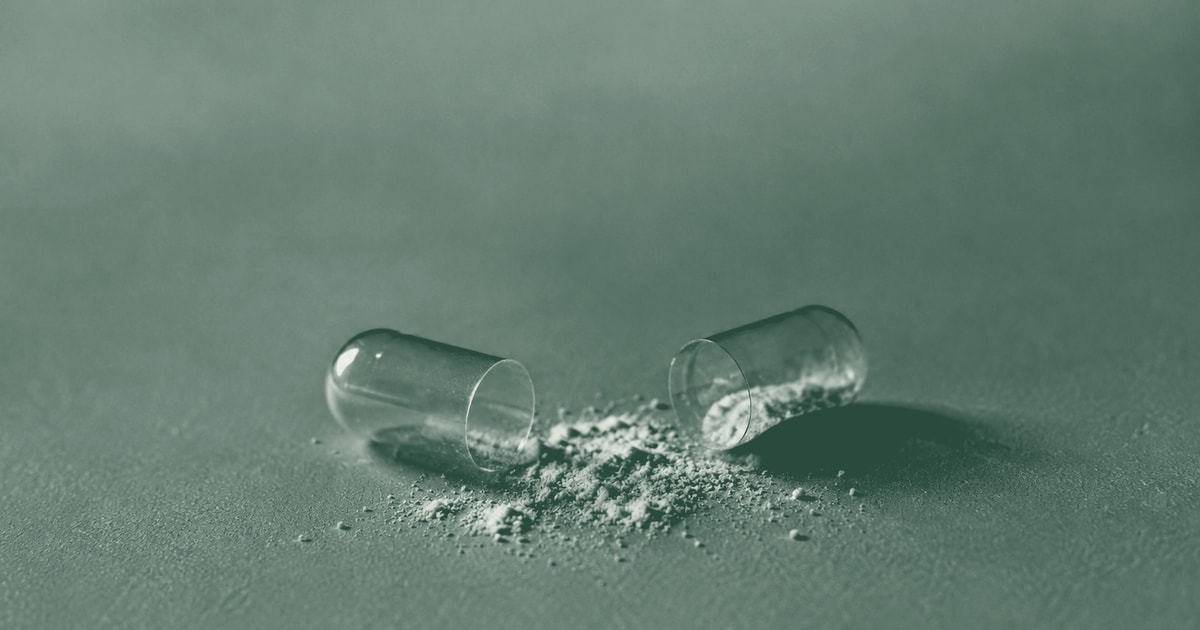 The two halves of a pulled-apart clear capsule lying on a plain surface with its white powder spilled between them, in a forest-green duotone.